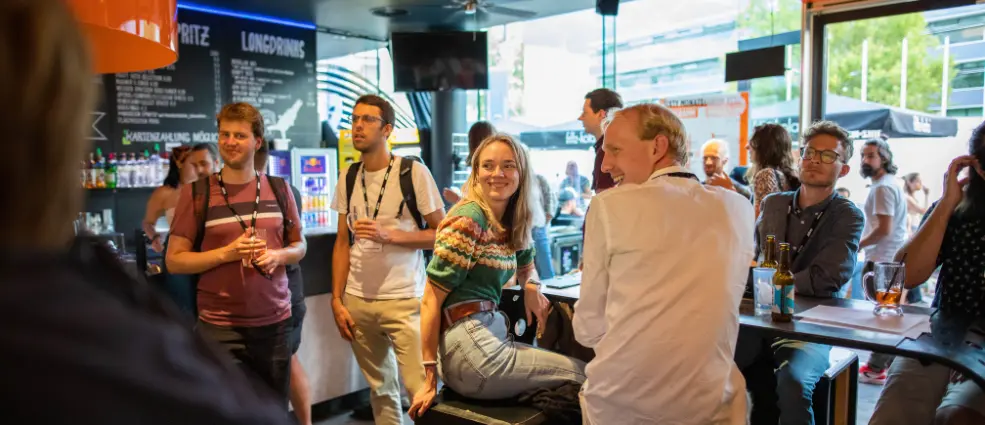 People wearing ECPR lanyards, socialising in a barv during an ECPR General Conference quiz night. A man in white laughs, a woman in a colourful jumper smiles.